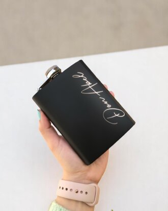Una mano sosteniendo un frasco negro con escritura cursiva, sobre una superficie blanca.